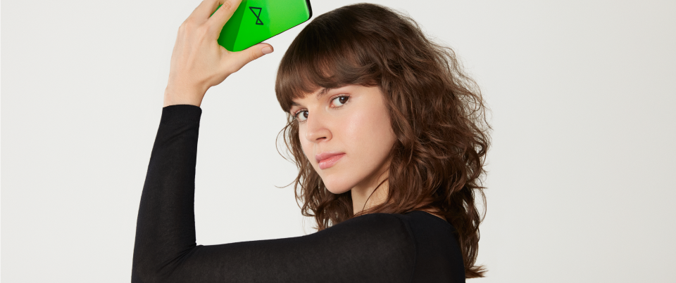 Uma mulher jovem segura um smartphone com uma mão acima da cabeça, onde vemos na tela um fundo verde neon e o logo do next. Ela tem cabelos cacheados castanhos e uma franja curta. Ela veste uma blusa de manga comprida preta.