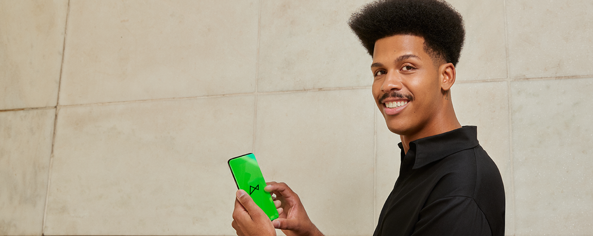 Vemos um homem negro, de cabelo crespo e bigode, sorrindo. Ele está vestindo uma camisa na cor preta, posicionado de lado, com a cabeça virada para olhar para câmera. Em suas mãos, vemos um celular com a tela verde, mostrando o logo do next.